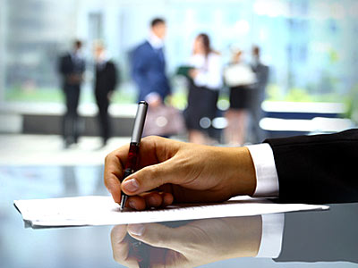 Eine Hand hält einen Stift und schreibt auf einem Dokument, im Hintergrund sind Menschen in formeller Kleidung.